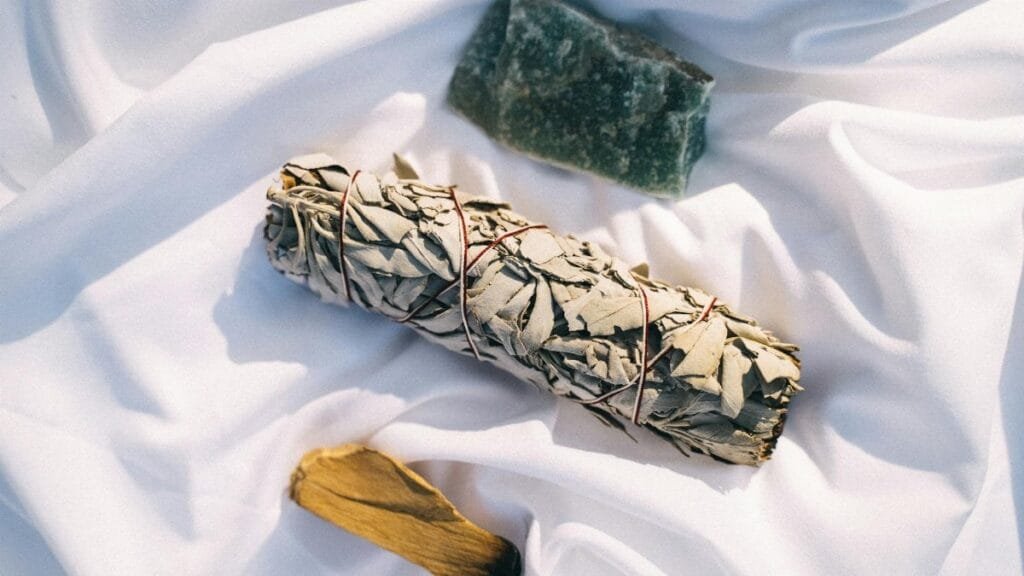A flat lay of sage, palo santo, and crystal on white fabric, symbolizing cleansing rituals.