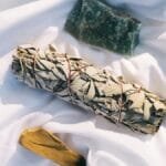 A flat lay of sage, palo santo, and crystal on white fabric, symbolizing cleansing rituals.