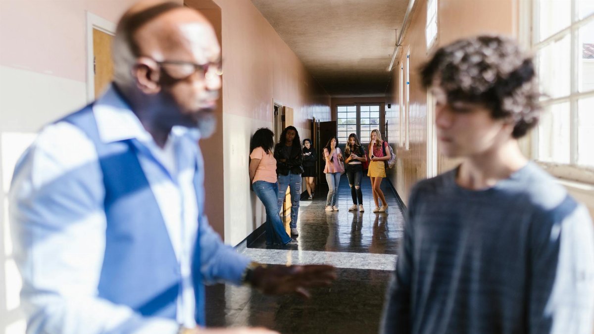 A blurred teacher talks to a student in a school hallway, with peers in the background.