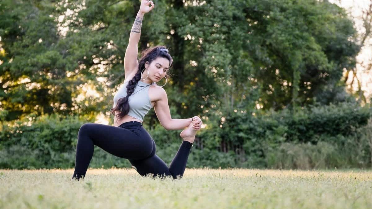 A young woman practicing a graceful yoga pose in a sunny field surrounded by lush greenery.