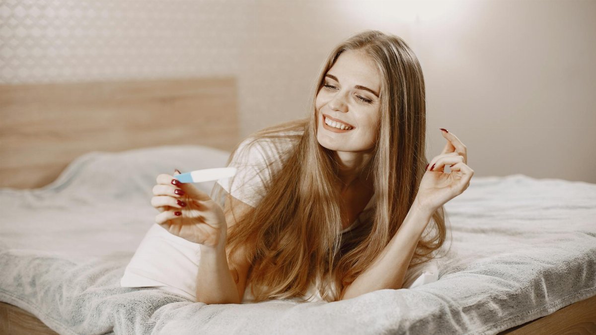 Smiling woman on bed holding a pregnancy test, expressing joy and anticipation.