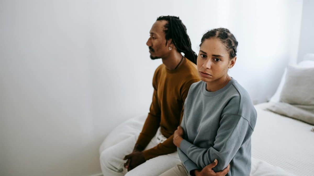 A couple sitting silently on a bed with tense expressions, suggesting conflict and emotional struggles.