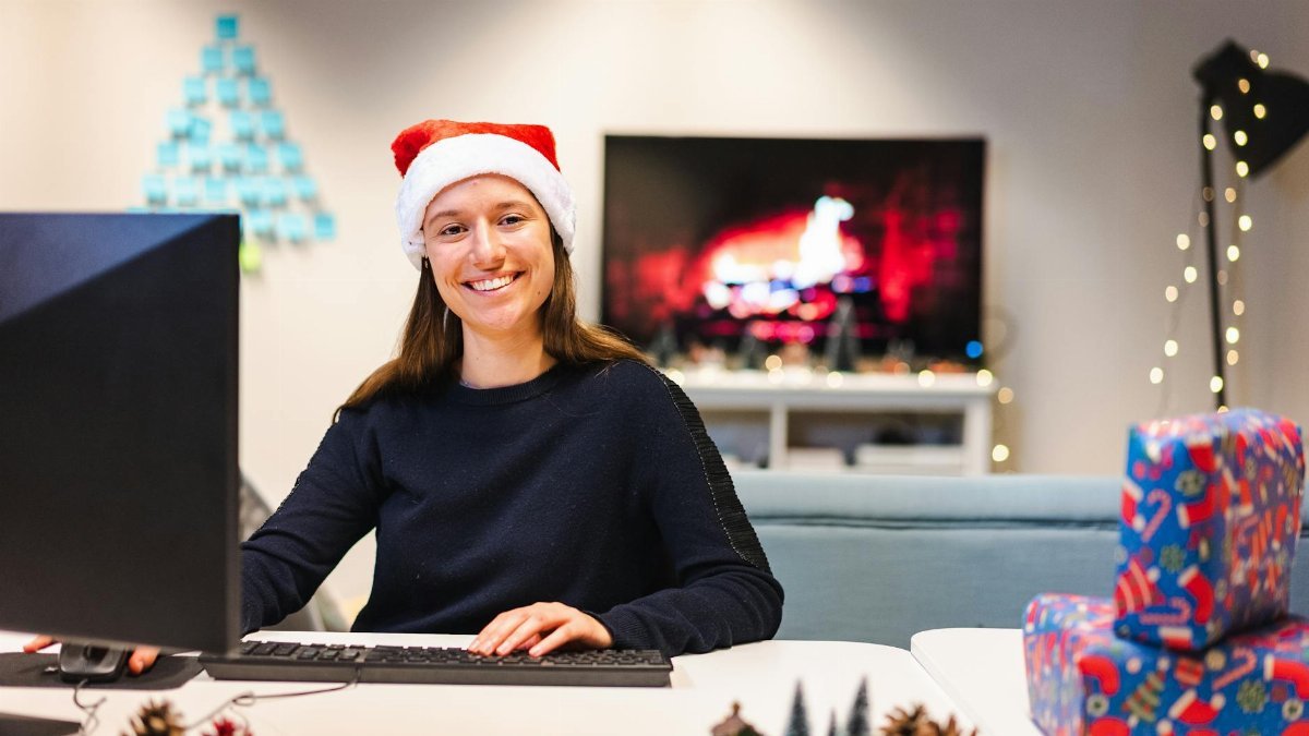 Smiling woman in Santa hat working on computer in festive office setting during Christmas season.