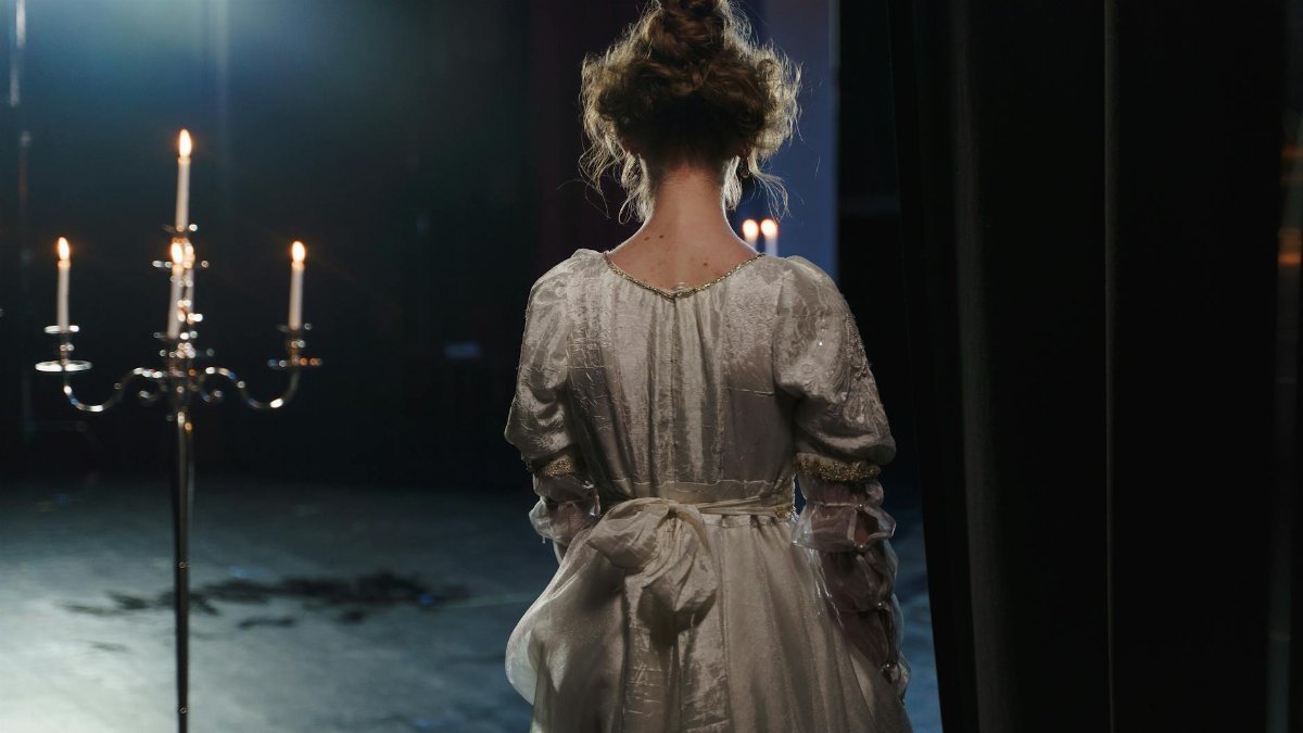 A woman in a white dress stands backstage under dramatic lighting, evoking a classic theatrical ambiance.