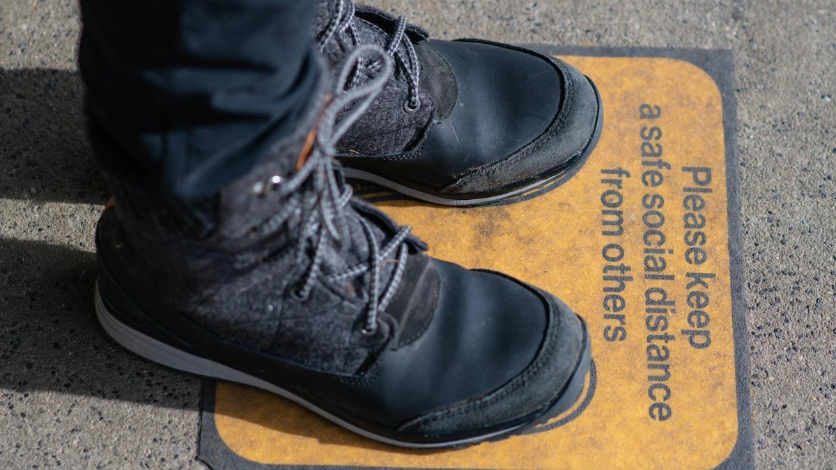 Black boots standing on a social distancing sign on pavement, emphasizing pandemic safety.