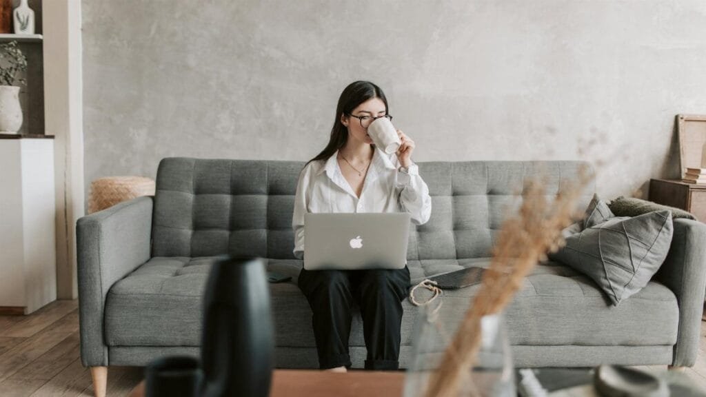 Woman sipping coffee while working on a laptop in a cozy home setting, highlighting remote work lifestyle.