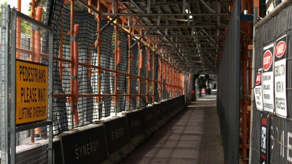 Pedestrian walkway through a construction site with safety signs and scaffolding overhead.
