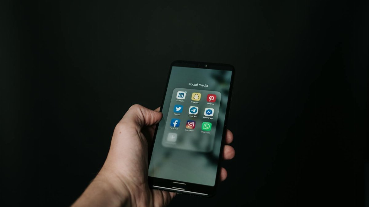 Close-up of a hand holding a smartphone displaying various social media app icons on a dark background.