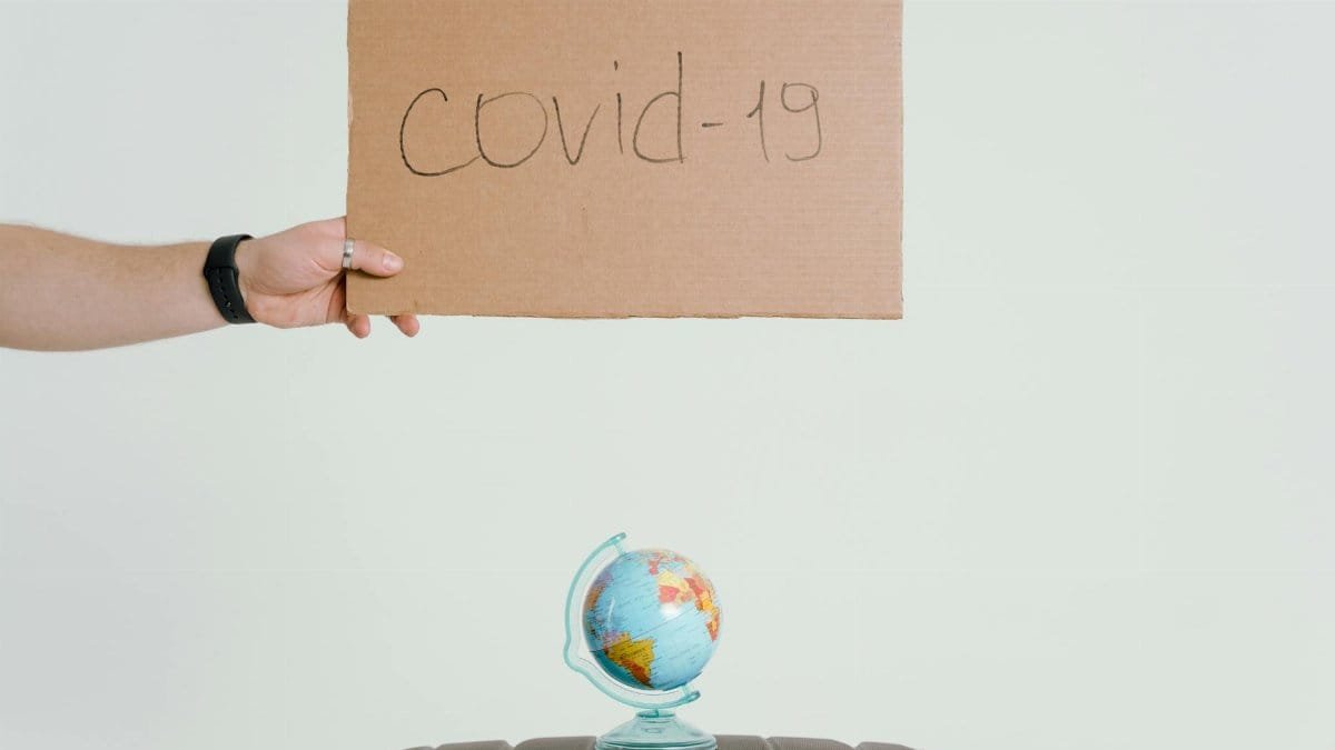 Hand holding covid-19 sign over globe and luggage, symbolizing pandemic travel impact.