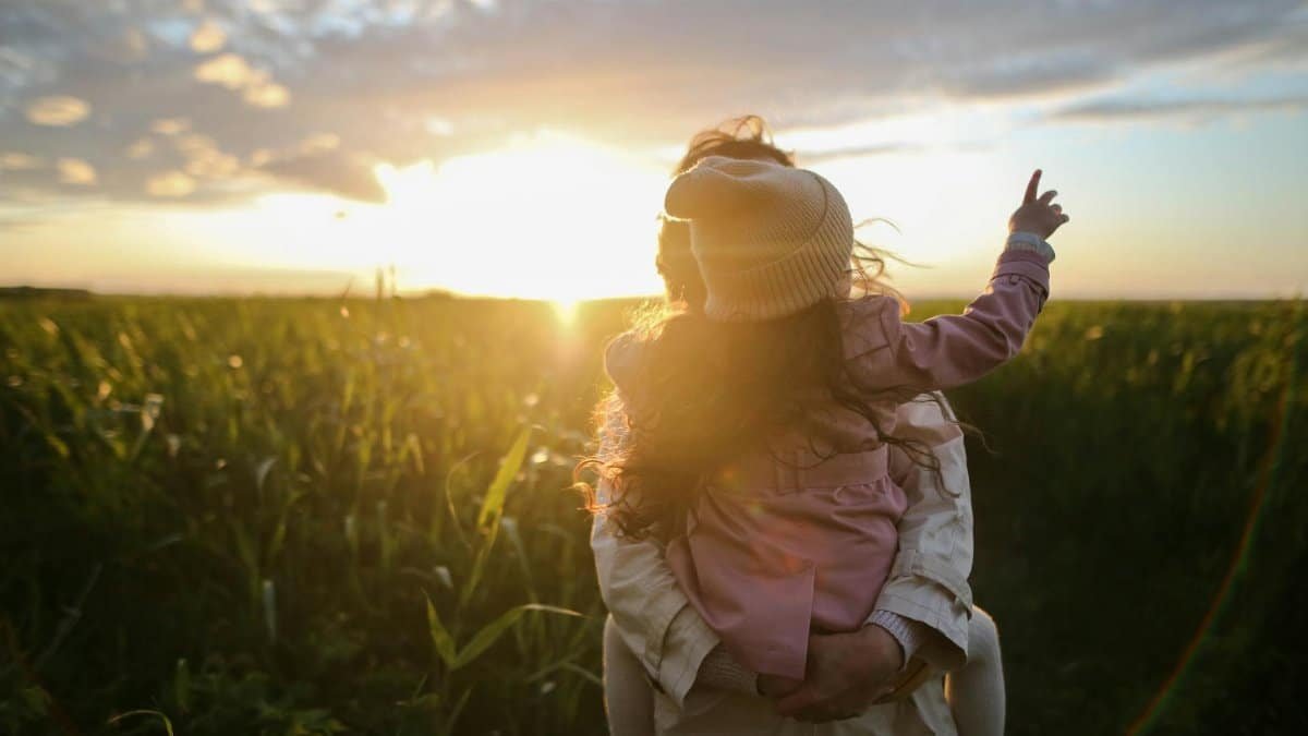 A mother and daughter embrace and point at the sunset in a grassy field.