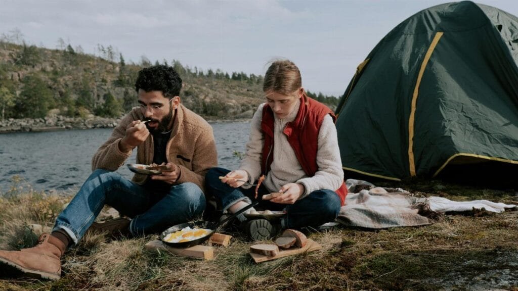 A couple sits by their tent enjoying a meal by a lakeside campsite during a day in nature.