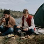A couple sits by their tent enjoying a meal by a lakeside campsite during a day in nature.