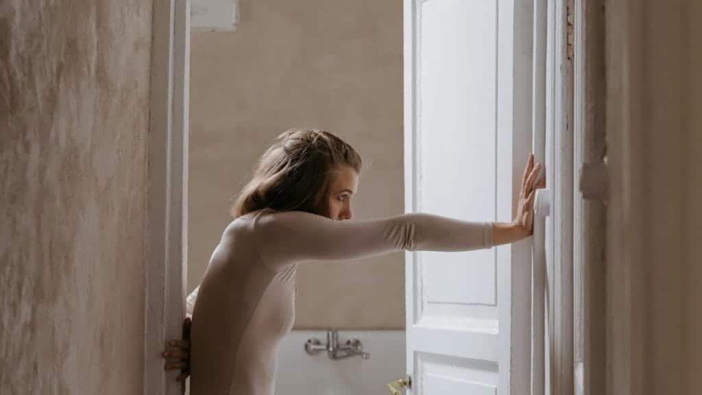 A woman leans on an open door in a rustic, minimalist indoor setting.