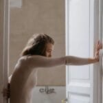 A woman leans on an open door in a rustic, minimalist indoor setting.