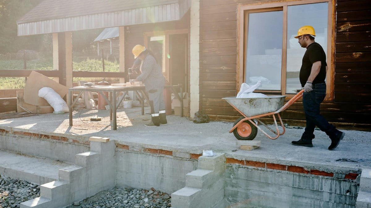 Two construction workers renovating a house exterior, carrying materials and working on woodwork.