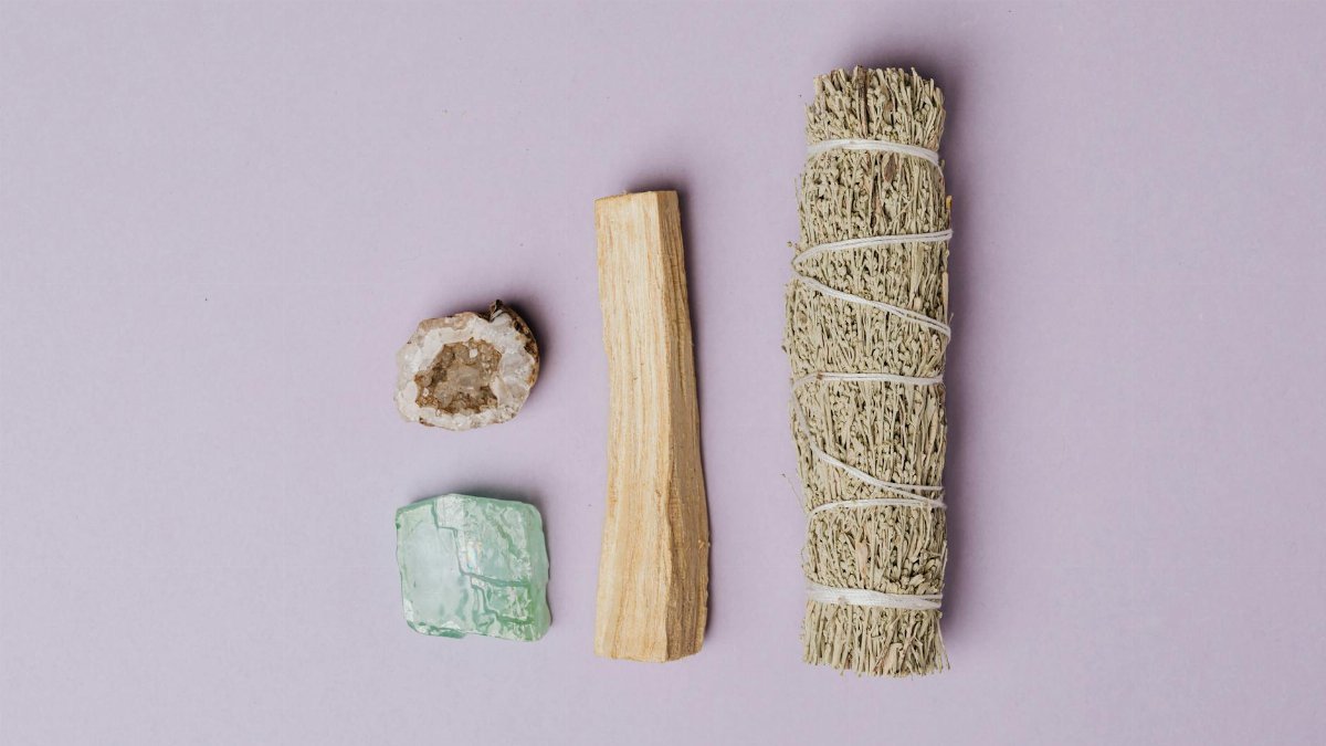 Flat lay of spiritual healing tools including sage, palo santo, quartz geode, and amazonite on a lavender background.