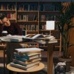 A young woman sits at a cluttered desk in a library, surrounded by books and a laptop.