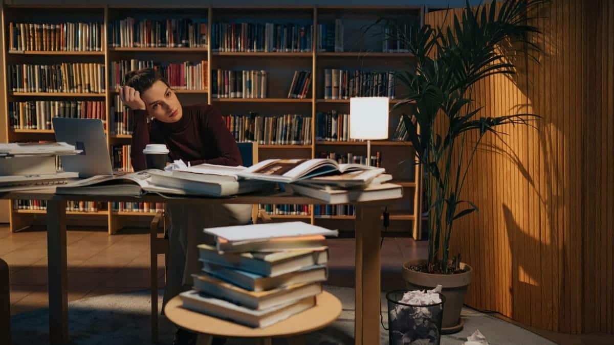 A young woman sits at a cluttered desk in a library, surrounded by books and a laptop.