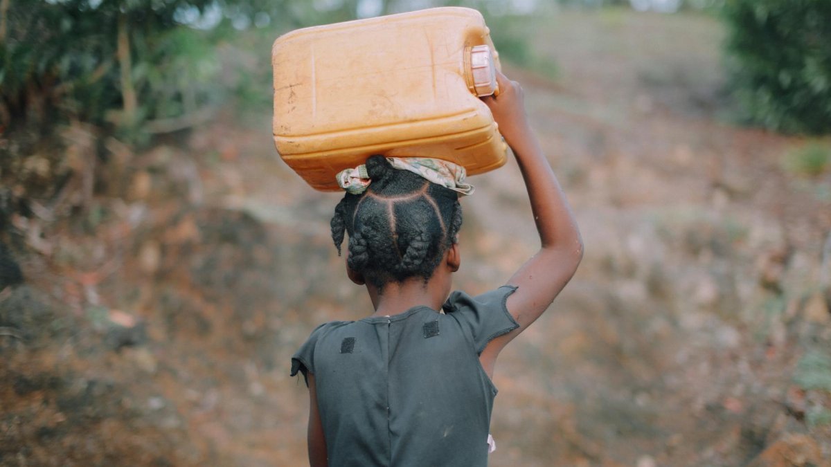 A young girl with braided hair carries a jug of water on her head in a rural area of Haiti.