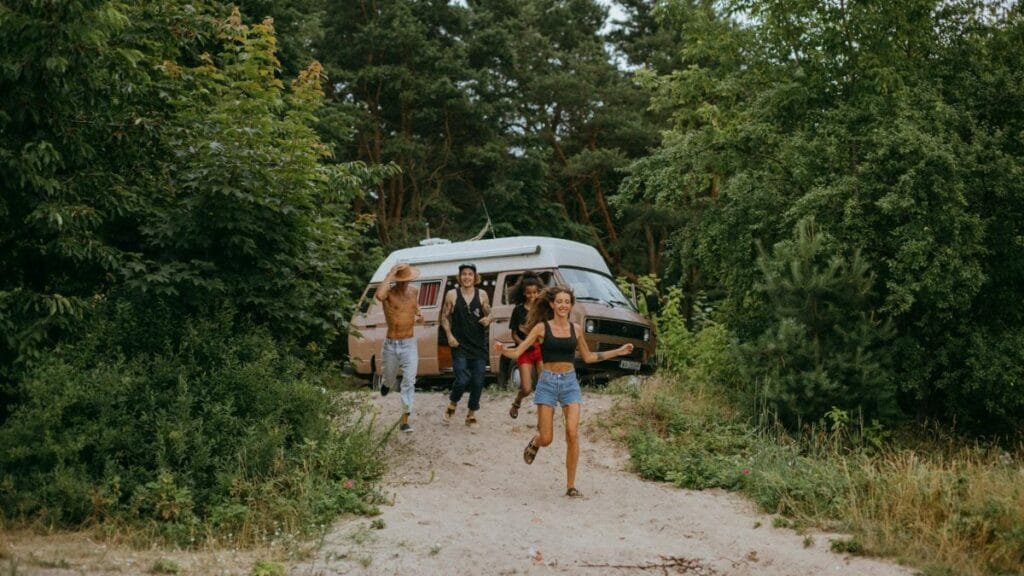 Group of friends running joyfully from a camper van in a lush forest setting during summer.