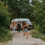 Group of friends running joyfully from a camper van in a lush forest setting during summer.