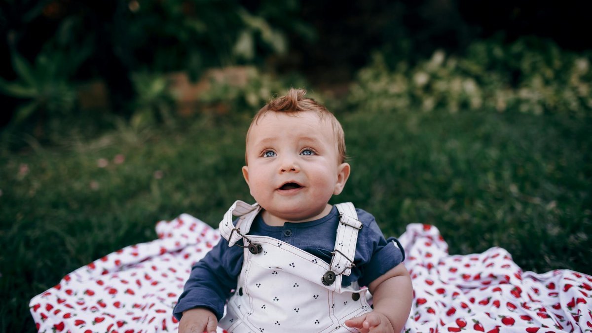 Cute baby with blue eyes sitting on a cherry-patterned blanket outdoors, enjoying a sunny day.