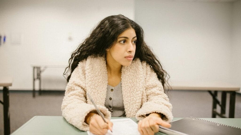 A college student writing on a test paper while looking away in a classroom setting.