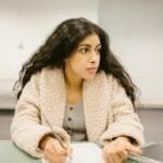 A college student writing on a test paper while looking away in a classroom setting.