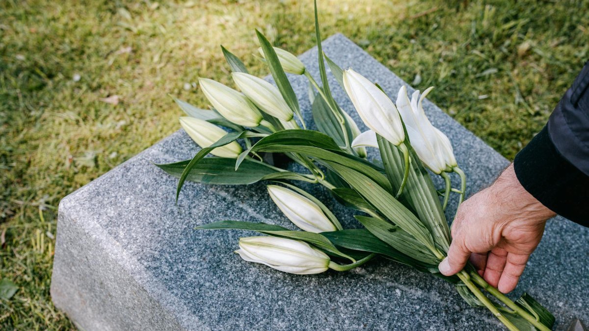 A hand placing white lilies on a granite tombstone in an outdoor cemetery.