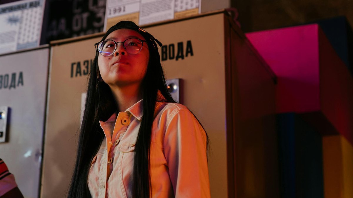 Teen girl in glasses stands thoughtfully near a vending machine, lit by colorful lights indoors.