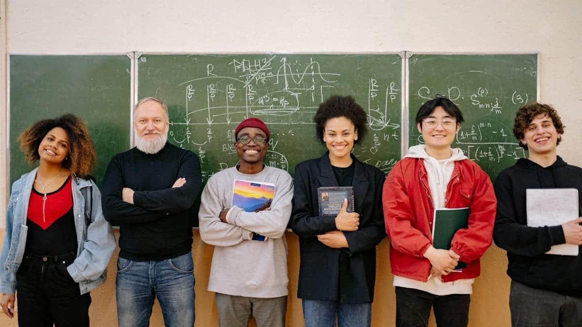 A diverse group of students and a teacher smiling in front of a chalkboard filled with formulas.