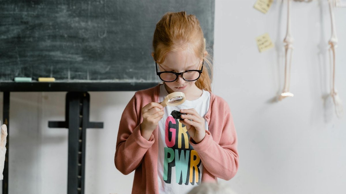A young girl in glasses focuses intently on an object with a magnifying glass, showcasing curiosity and exploration.