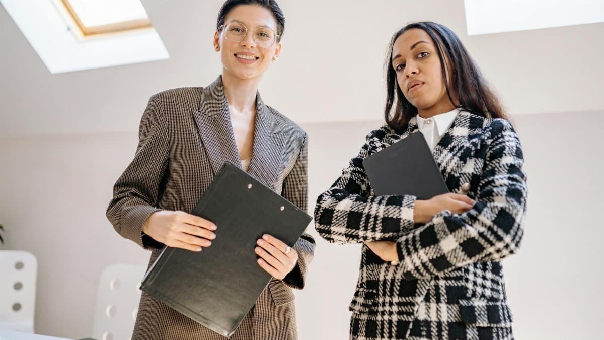 Two confident businesswomen in stylish suits holding folders in a modern office setting.