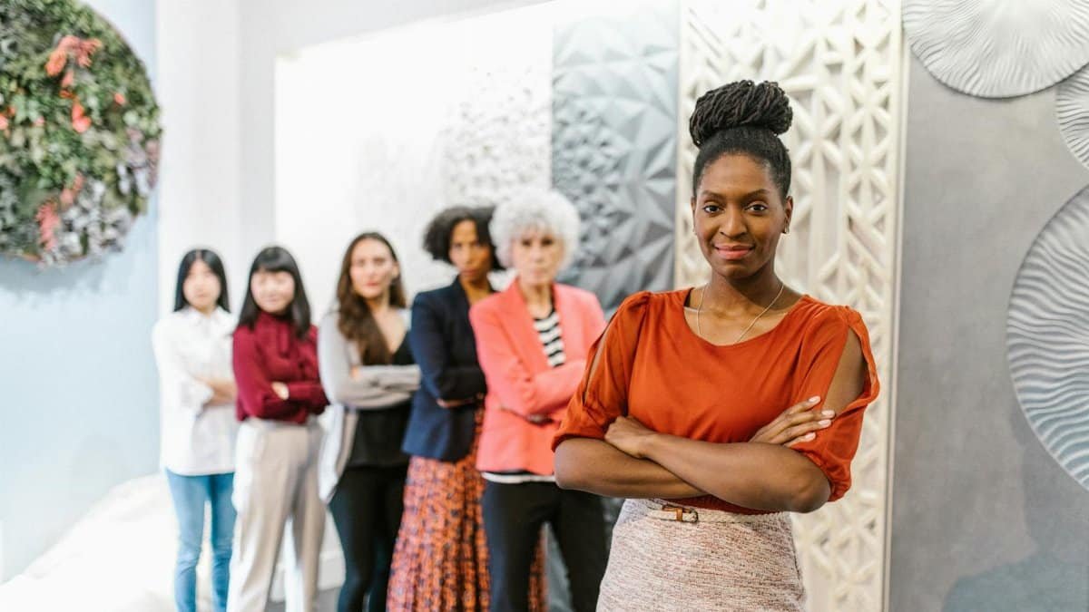 A confident female leader stands with diverse female colleagues in a modern office setting.