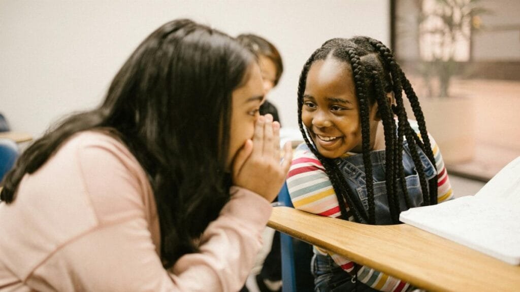 Two girls whisper and smile in a classroom, fostering friendship and joy.