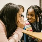 Two girls whisper and smile in a classroom, fostering friendship and joy.