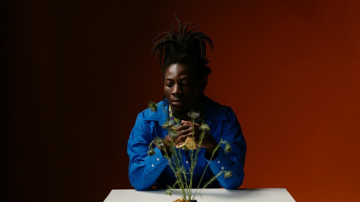 Modern fashion portrait of a man in blue with unique hair, posing with plants on a table.