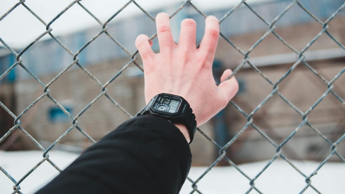 A hand wearing a digital watch reaching through a chain link fence in an outdoor setting.