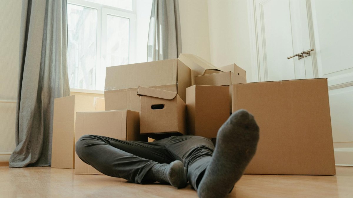 A person lies down tired among cardboard boxes in a bright room, depicting moving day stress.