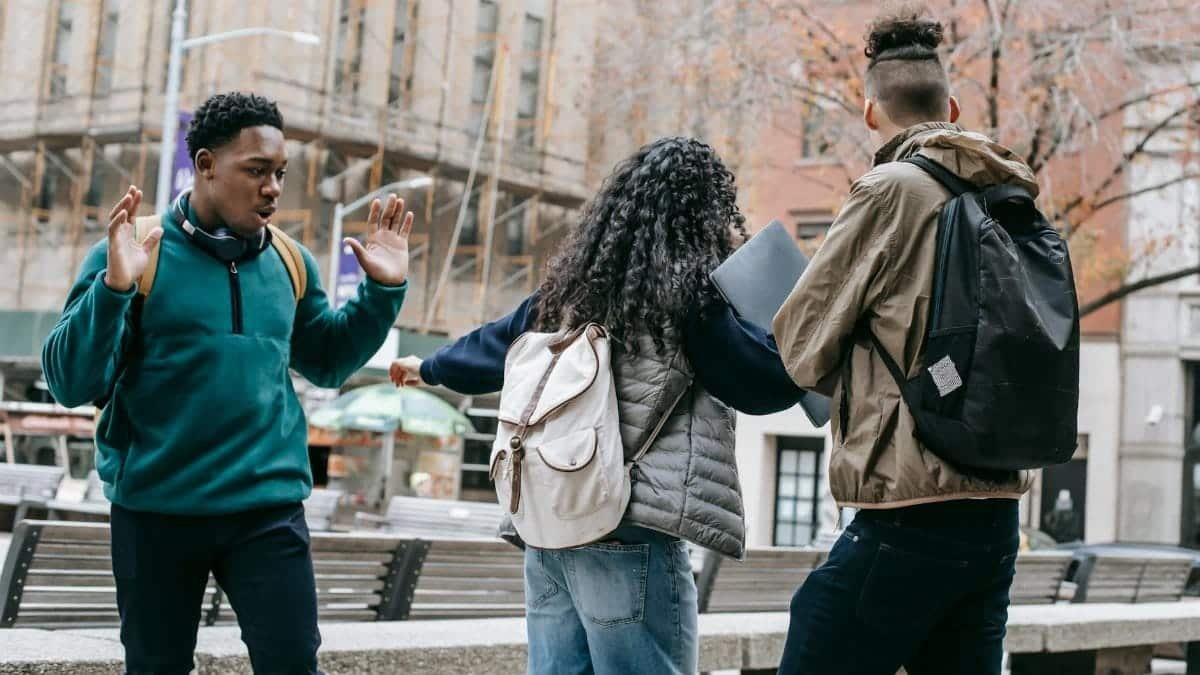 Back view of faceless student with backpack pushing away diverse classmates while standing in city park