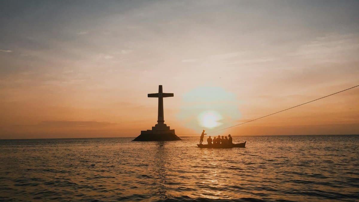 A striking silhouette of a cross and boat against a vibrant sunset over the sea.