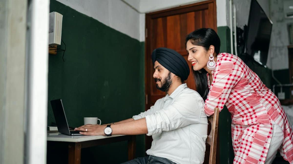 Side view of positive young Sikh man in shirt and turban working on laptop at home while wife leaning on chair behind