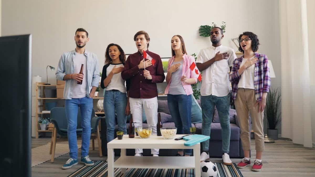 A diverse group of friends indoors, singing along to the national anthem while watching a TV broadcast.