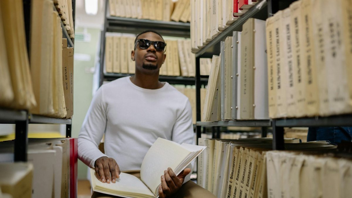 African American man with visual impairment reading braille book in a library.