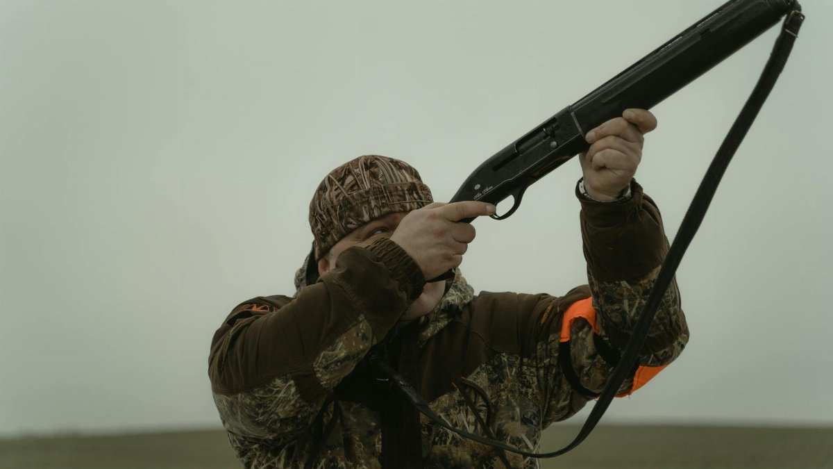 Hunter wearing camouflage gear aiming a shotgun in an open field setting, demonstrating outdoor hunting activity.