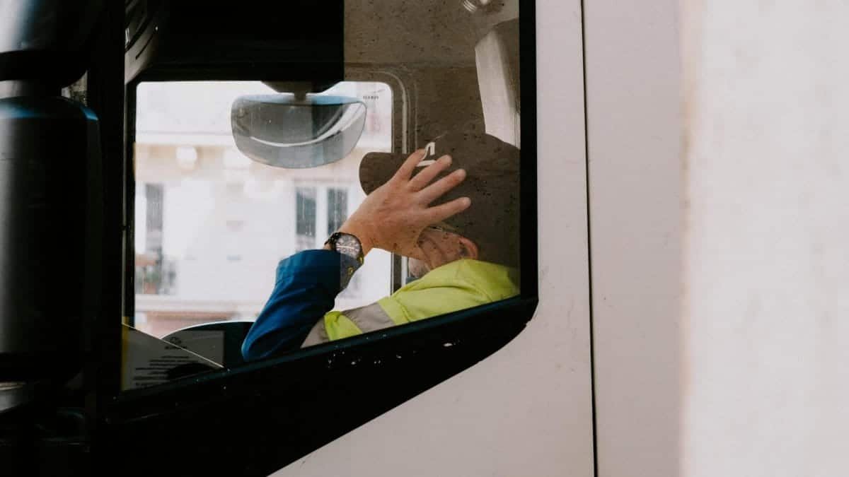 A truck driver in neon jacket rests with hand on head inside vehicle cabin, view from the side window.