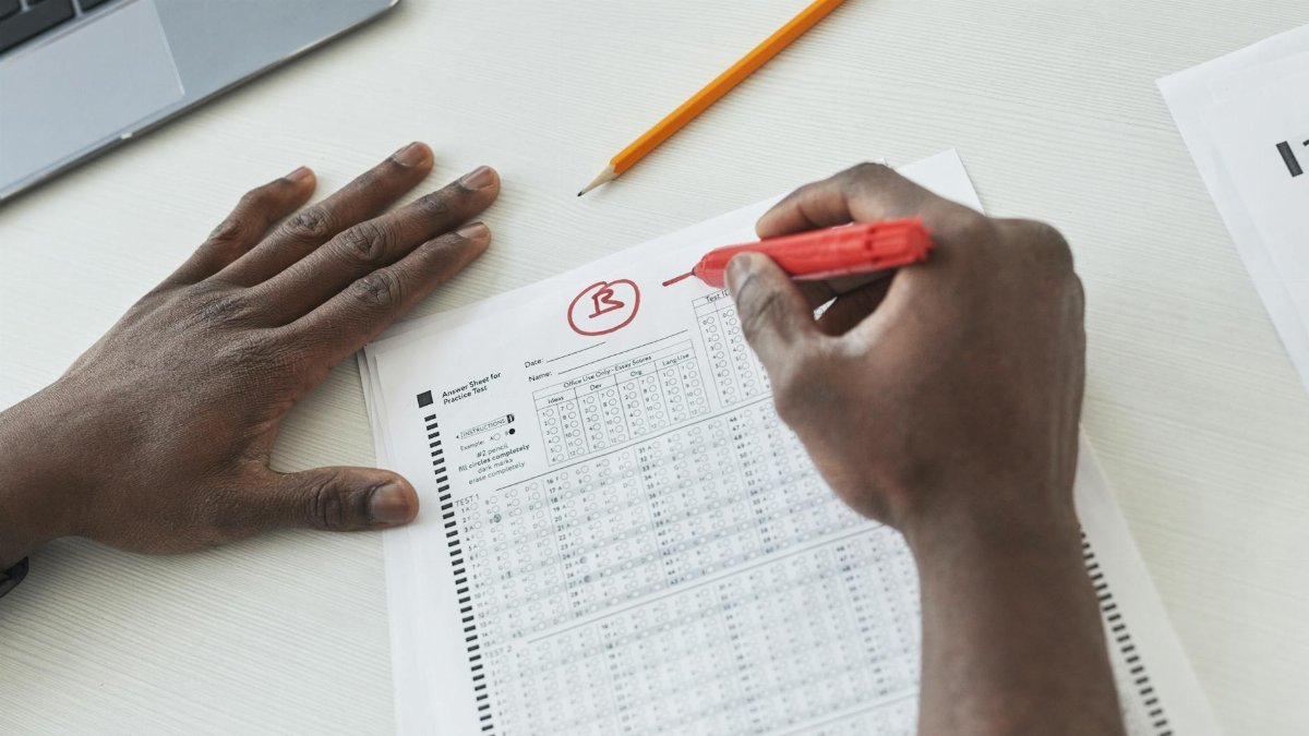 Close-up of a teacher grading multiple choice exams with a red pen on a desk.