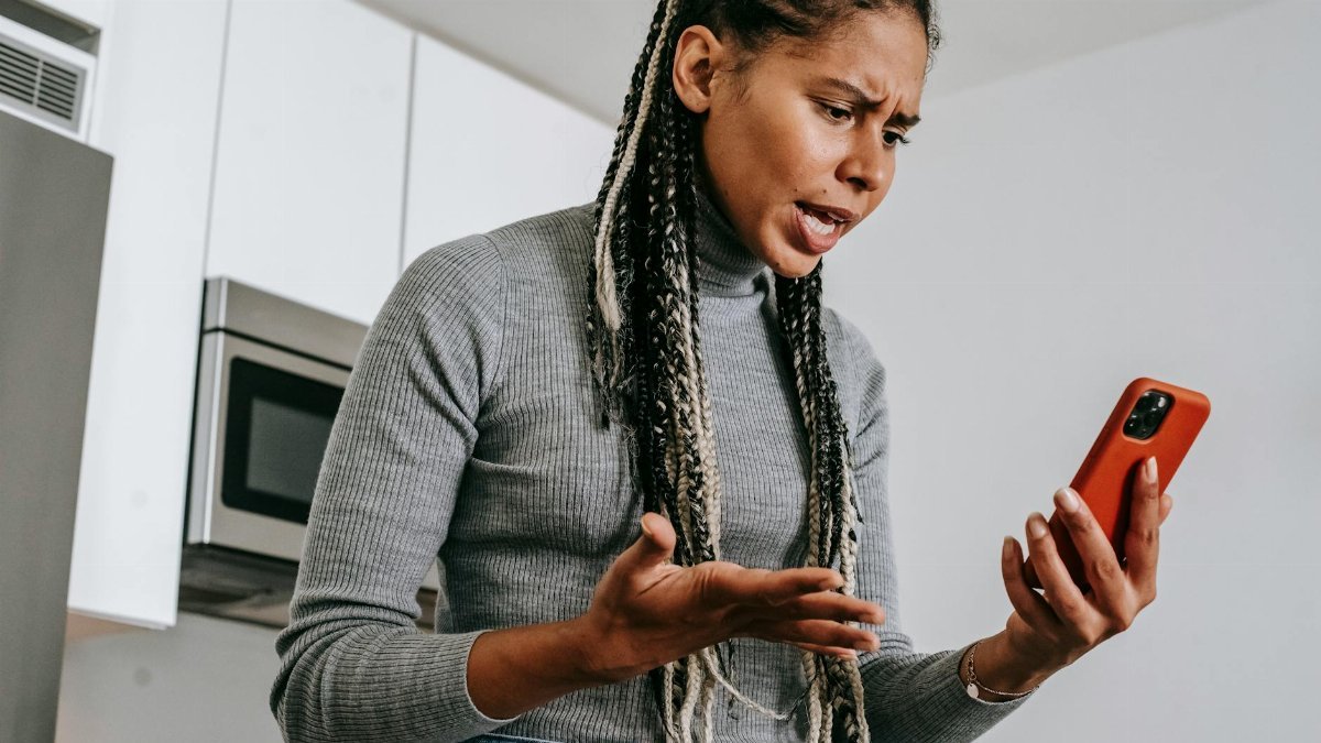 A young woman expressing frustration during a phone call in her kitchen, using a smartphone.