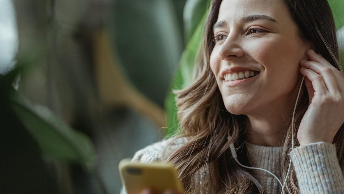 Happy young woman with long wavy hair smiling and adjusting earphones while listening to music via mobile phone