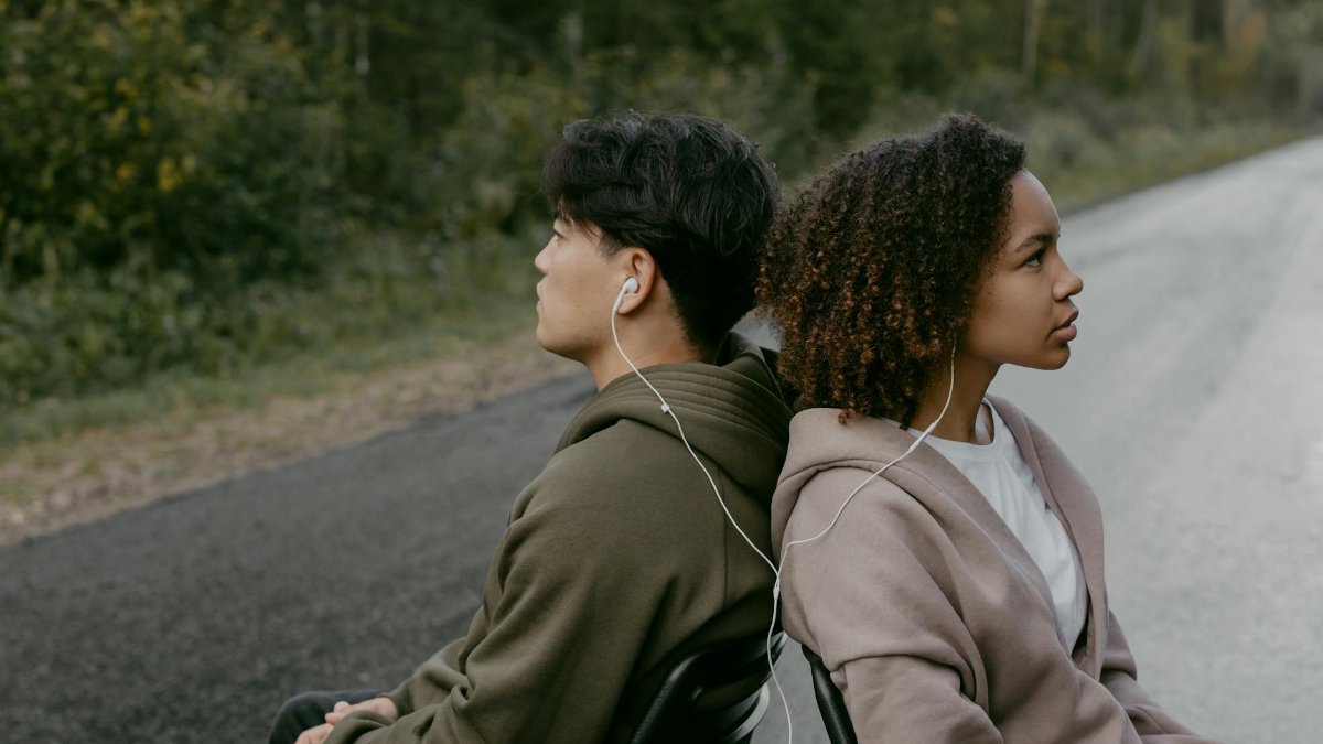 Two young adults sitting back to back, sharing headphones on a country road.
